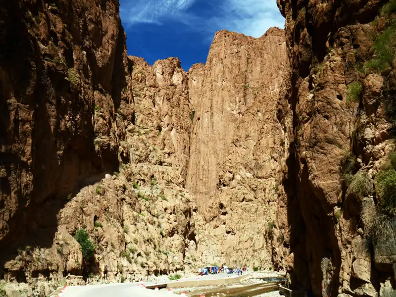Die stemmige rote Felswand der Todra Schlucht ragen hoch empor, mit Menschen am Flussbett.