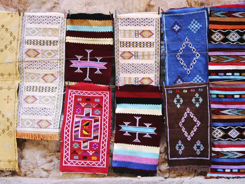 Auswahl an traditionellen Teppichen mit unterschiedlichen Farben und Mustern, die an einer Steinwand hängen.