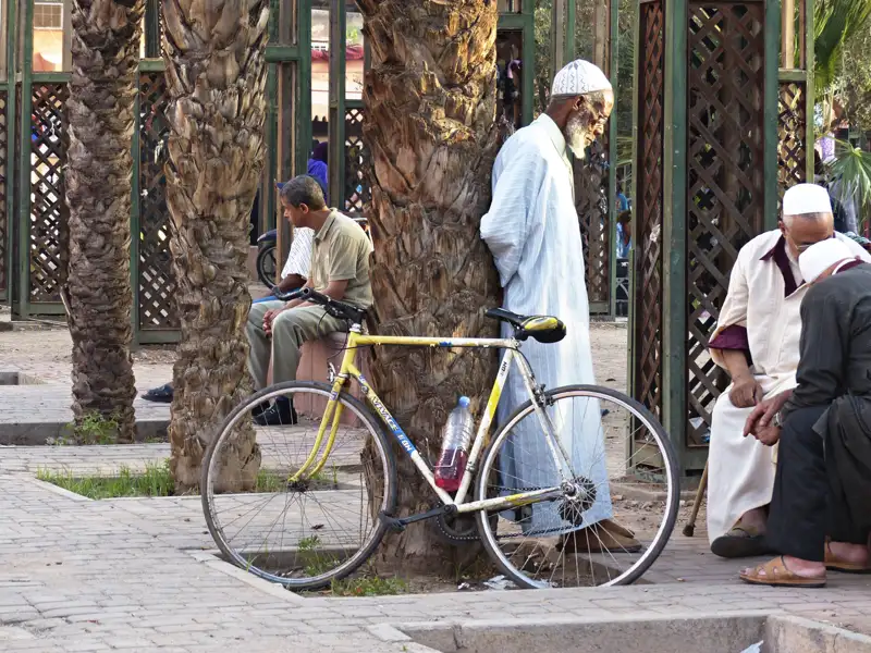 Männer in traditioneller Kleidung ruhen sich in einem Park in Marrakesch aus. Ein gelbes Fahrrad steht an einem Baum gelehnt.