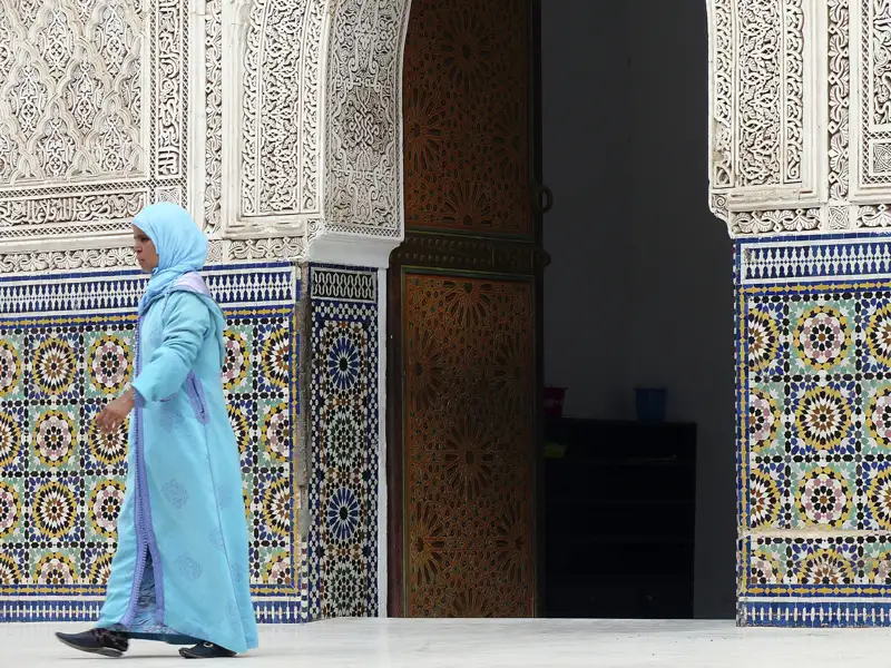 Frau in traditioneller Kleidung vor verzierter Wand mit Mosaikfliesen.