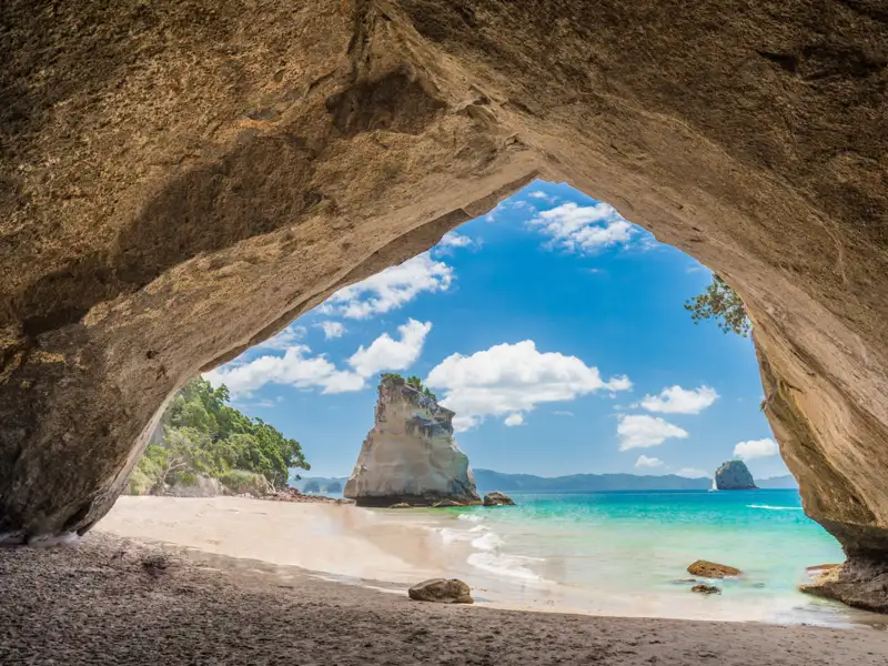 Blick durch eine Meereshöhle auf einen Sandstrand mit einzigartigen Felsformationen im türkisfarbenen Wasser.