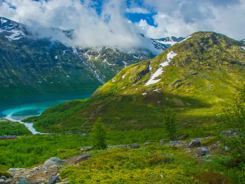 Türkisfarbener See in einem Tal, umgeben von grünen Bergen mit Schneefeldern.