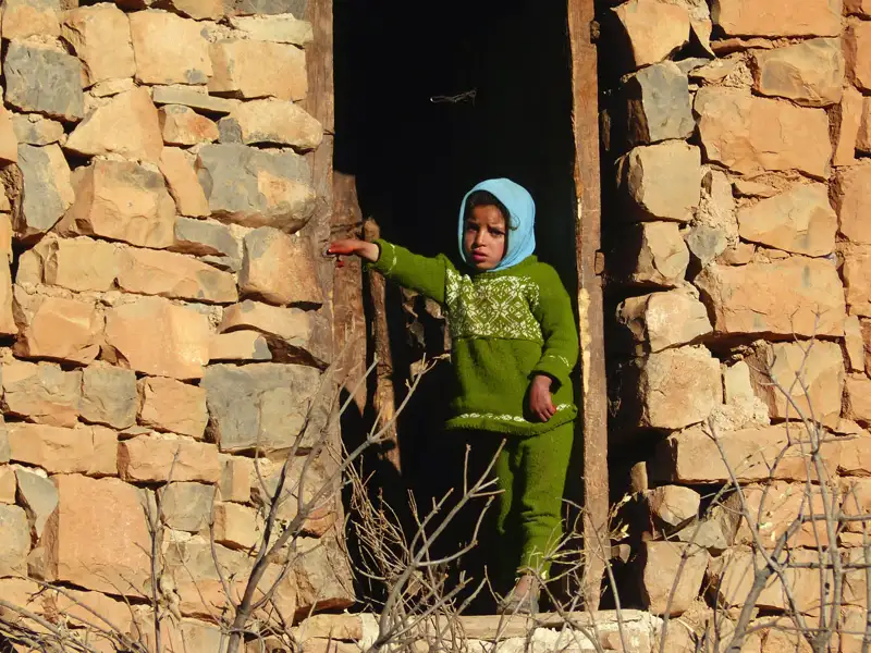 Kind in grüner Kleidung und blauem Kopftuch im Türrahmen eines Steinhauses.