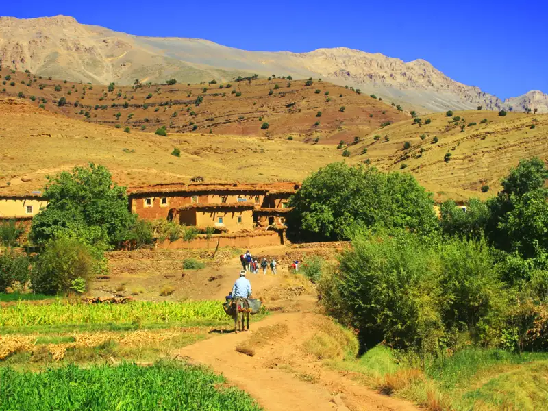 Wanderer auf einem Bergpfad in Marokko, mit einem traditionellen Dorf und Bergen im Hintergrund.