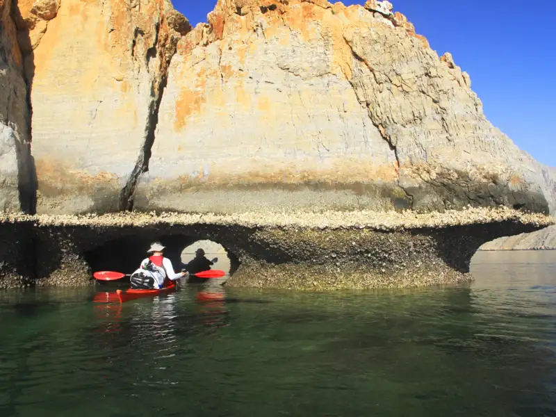Kajakfahrer paddeln unter einem natürlichen Felsbogen an der Küste entlang.