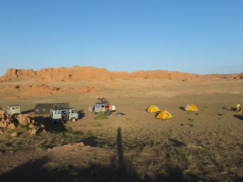 Campingplatz in der Wüste mit Zelten und Fahrzeugen, vor dem Hintergrund roter Felsformationen.