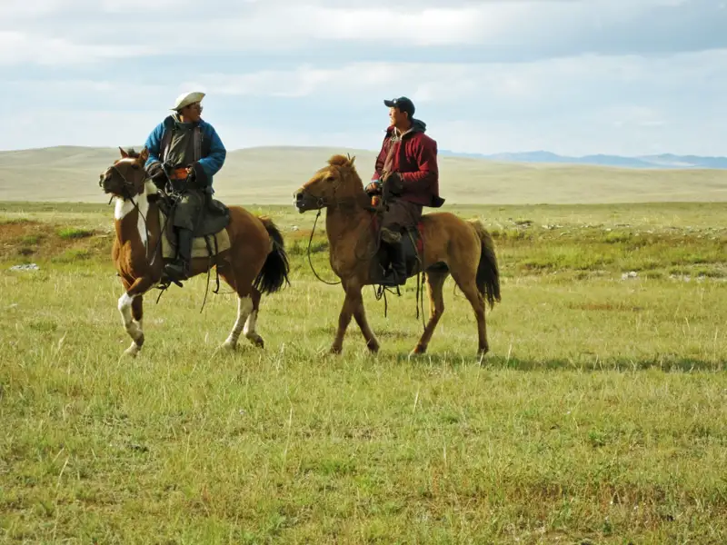 Zwei Reiter in traditioneller Kleidung durchqueren die Steppe auf ihren Pferden.