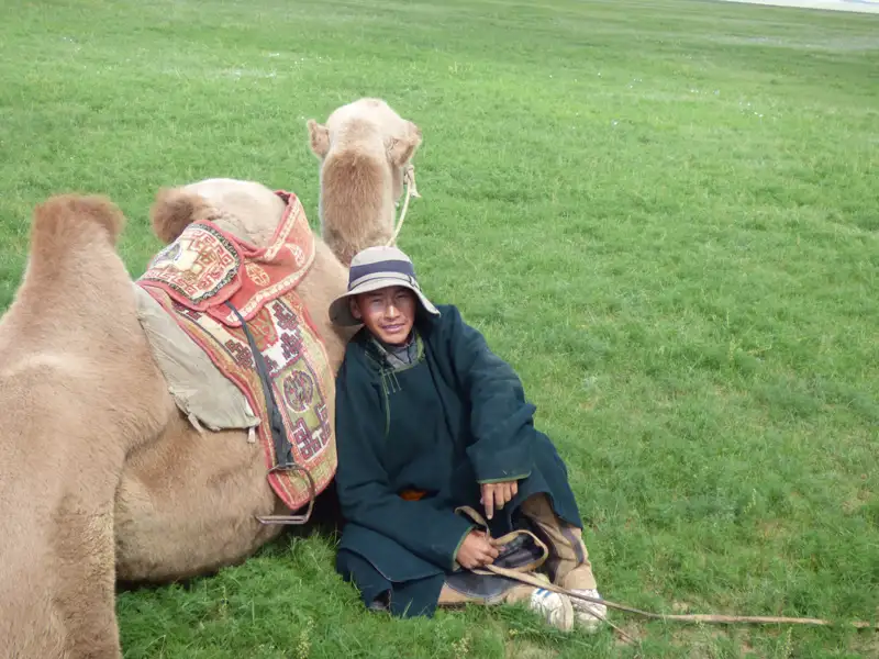 Reisender in traditioneller Kleidung pausiert neben einem Kamel auf einer grünen Wiese.
