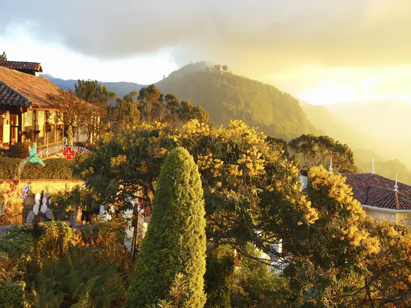 Sonnenuntergang über einer Bergkette, aufgenommen von einer Unterkunft mit Ziegeldach und umgebender Vegetation.
