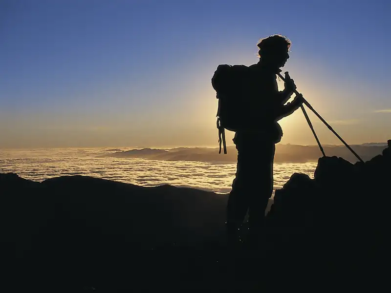Silhouette eines Wanderers mit Rucksack und Wanderstöcken, der auf einem Berggipfel steht und den Sonnenaufgang über einem Wolkenmeer beobachtet.