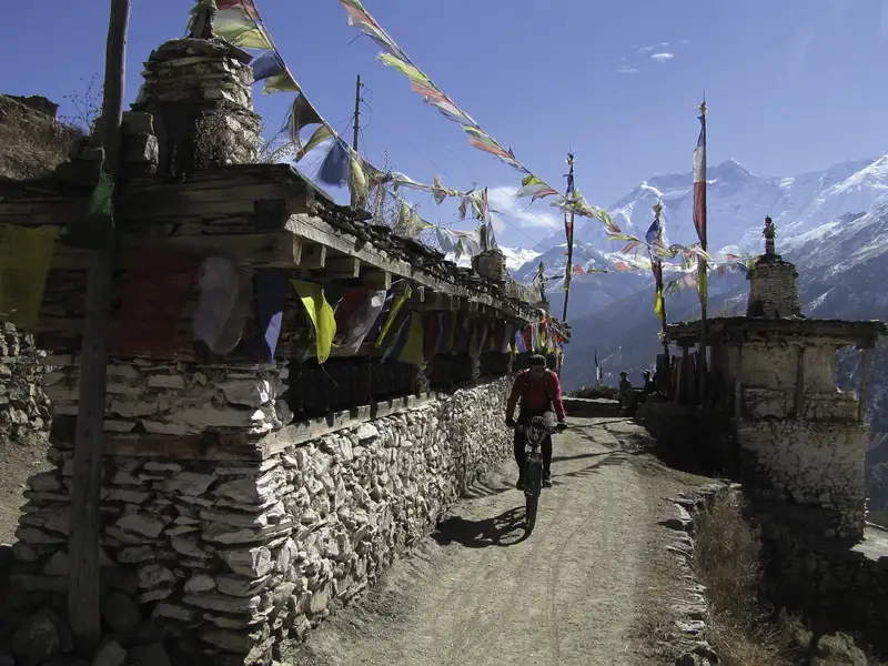 Radfahrer auf einem Bergpfad im Himalaya, vorbei an traditionellen Steinbauten mit Gebetsfahnen und schneebedeckten Bergen im Hintergrund.