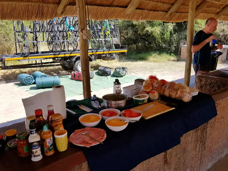 Frühstücksbuffet mit verschiedenen Zutaten und Fahrrädern auf einem Anhänger im Hintergrund, bereit für eine Radtour.
