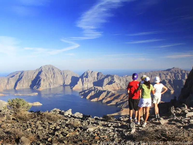 Drei Personen stehen auf einem Berg und blicken auf eine Bucht und die umliegenden Berge.