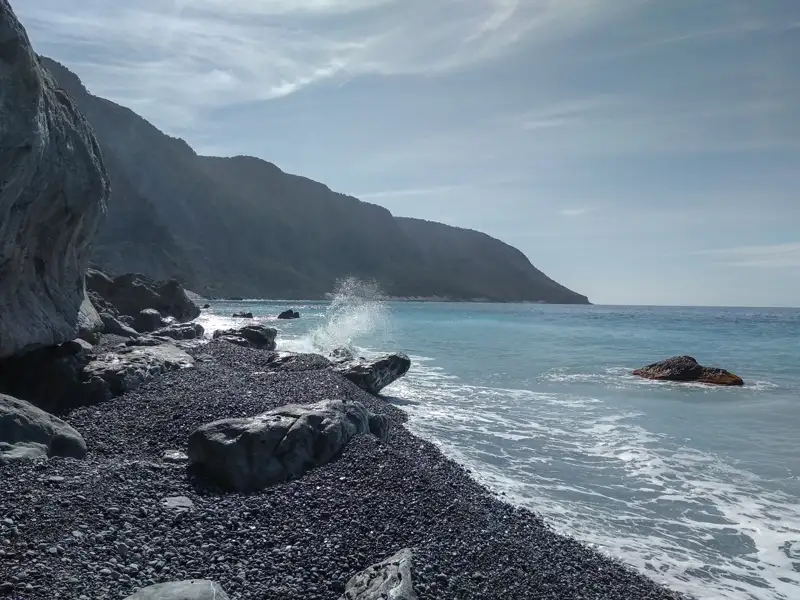 Wellen, die auf einen Kieselstrand treffen, mit einer Bergkette im Hintergrund.