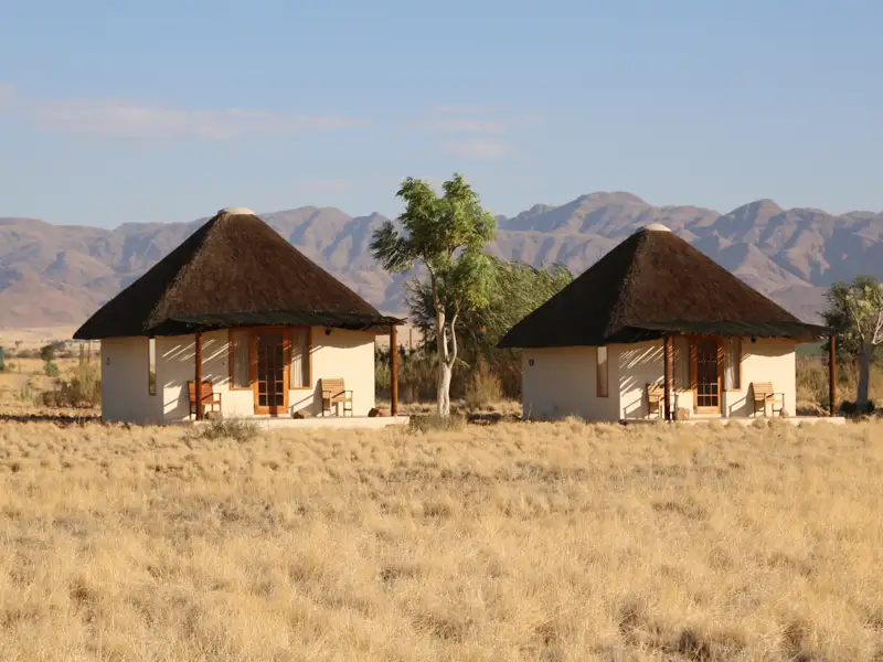 Unterkünfte der Reise: Traditionelle Rundhütten mit Strohdächern und Veranden in einer trockenen Landschaft mit Bergen im Hintergrund.