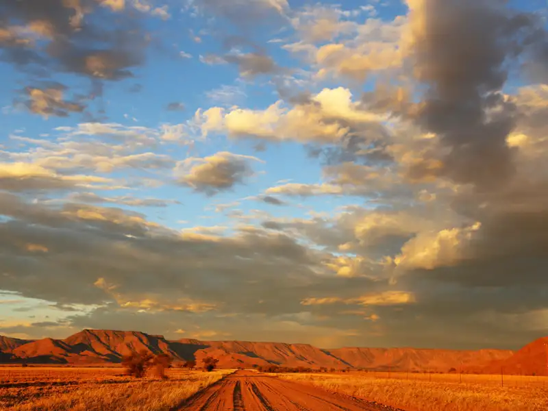 Unbefestigte Straße in einer trockenen Landschaft mit Bergen im Hintergrund bei Sonnenuntergang.