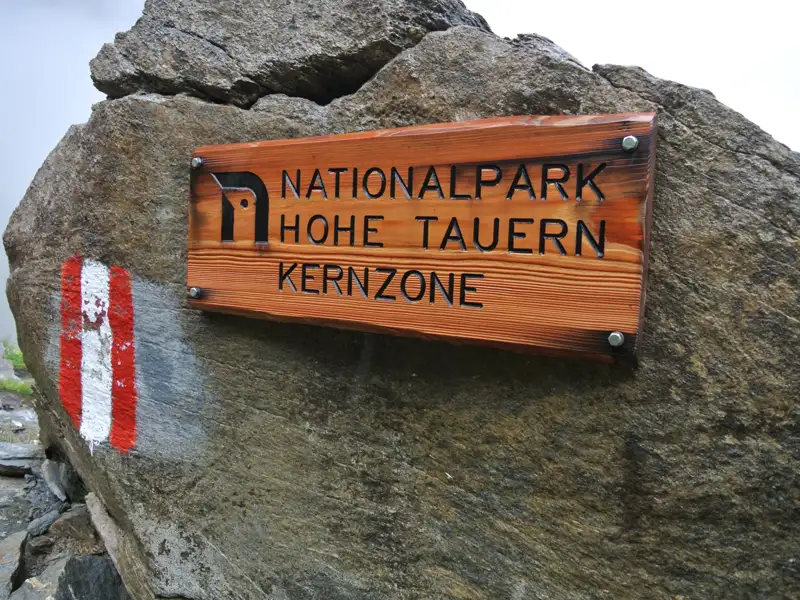 Schild mit der Aufschrift 'Nationalpark Hohe Tauern Kernzone' und rot-weiße Wegmarkierung an einem Felsen.