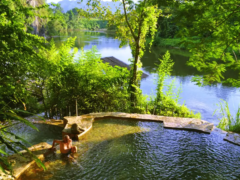 Reisender entspannt in einem natürlichen Flussbad mit Blick auf den Fluss und die umliegende Natur.