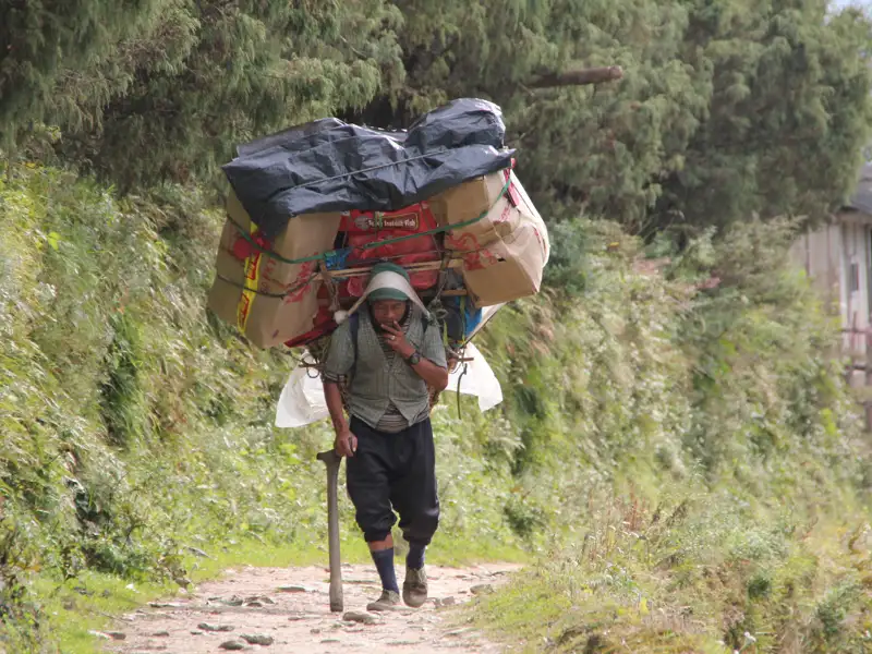 Ein Träger trägt eine schwere Last auf seinem Rücken, während er einen Bergpfad entlang wandert.