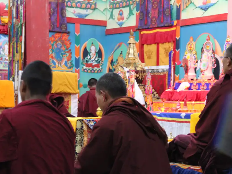 Mönche in roten Roben nehmen an einer Zeremonie in einem Tempel teil, der mit bunten Wandmalereien und religiösen Symbolen geschmückt ist.