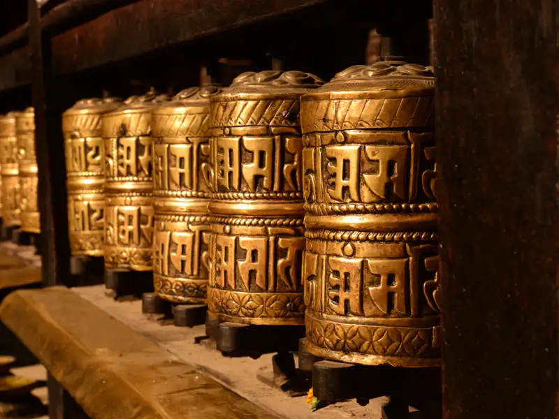 Goldene Gebetsmühlen mit tibetischen Inschriften, die in einer Gebetsstätte aneinandergereiht sind.
