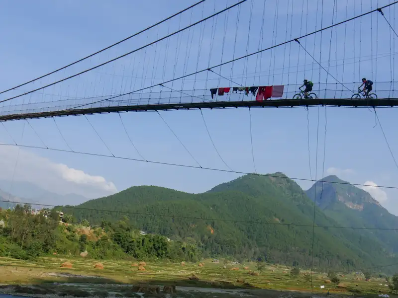 Zwei Radfahrer auf einer Hängebrücke mit Bergen im Hintergrund.