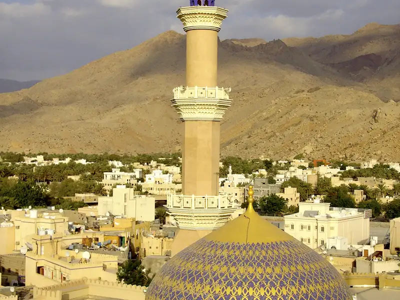 Blick auf die goldene Kuppel und das Minarett einer Moschee, eingebettet in eine Ortschaft mit Palmen und Bergen im Hintergrund.