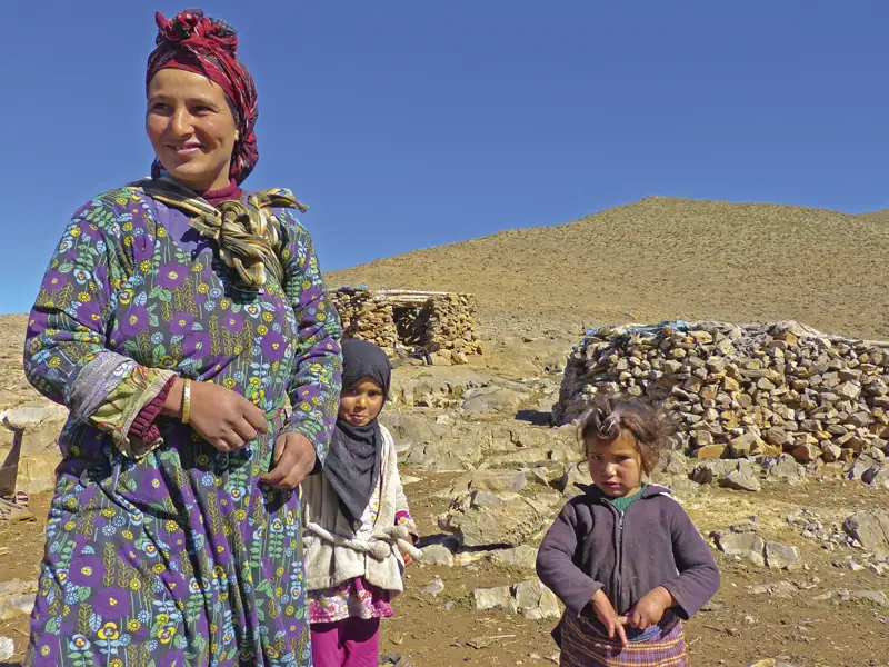 Eine Frau mit zwei Kindern vor traditionellen Steinhütten in einer bergigen Landschaft.