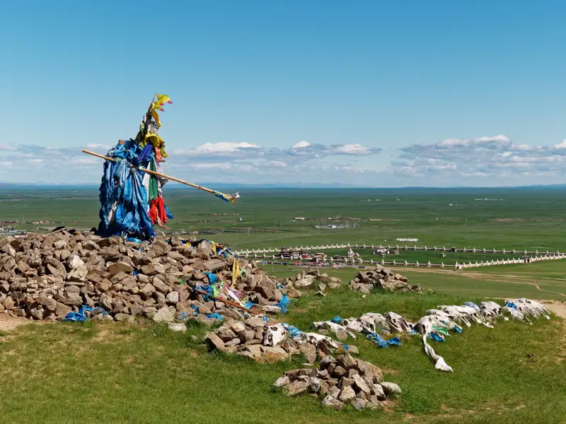Gebetsfahnen und eine Gebetsstange auf einem Steinhaufen in der Mongolei. Im Hintergrund ist die weite Landschaft der Steppe und eine ummauerte Anlage zu sehen.