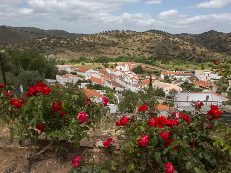 Blick auf ein weißes Dorf inmitten einer hügeligen Landschaft, eingerahmt von blühenden roten Rosen.