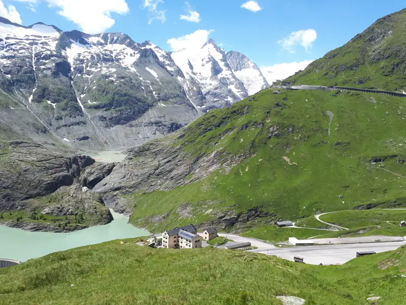 Stausee in den Alpen umgeben von schneebedeckten Bergen und einer Bergstraße.
