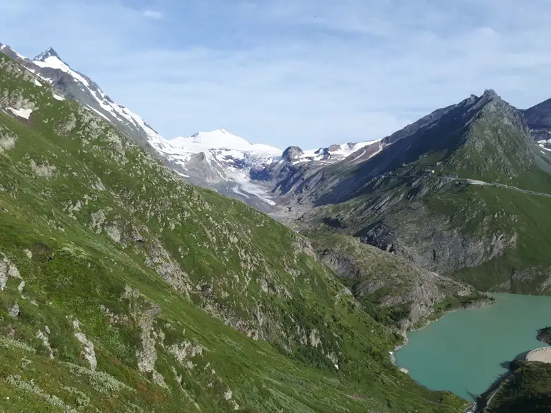Türkisfarbener Stausee in den Alpen, umgeben von grünen Berghängen und schneebedeckten Gipfeln.