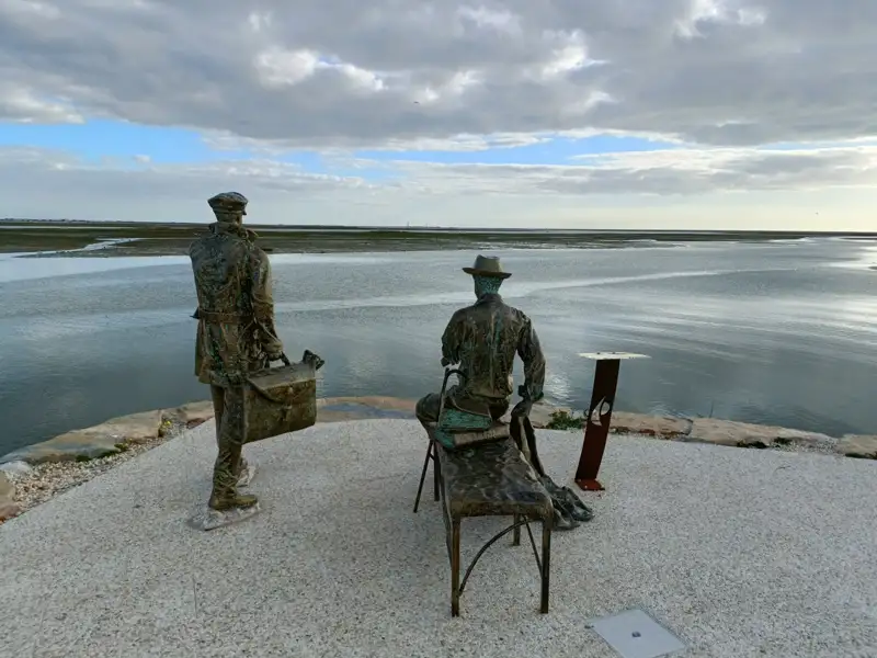 Zwei Statuen am Ufer eines Gewässers. Die stehende Figur trägt einen Koffer, die sitzende Figur ruht auf einer Bank. Beide blicken in die Ferne über das Wasser.