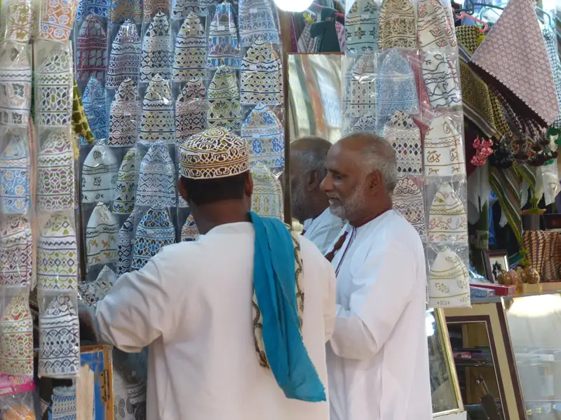 Zwei Männer begutachten traditionelle Kopfbedeckungen an einem Marktstand. Die Hüte sind in Plastik verpackt und zeigen verschiedene Muster und Farben.