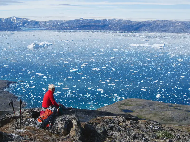 Ein orangefarbenes Boot, bereit für eine Exkursion in der Arktis, liegt an einem felsigen Ufer. Im Hintergrund spiegeln sich schneebedeckte Berge im Wasser.