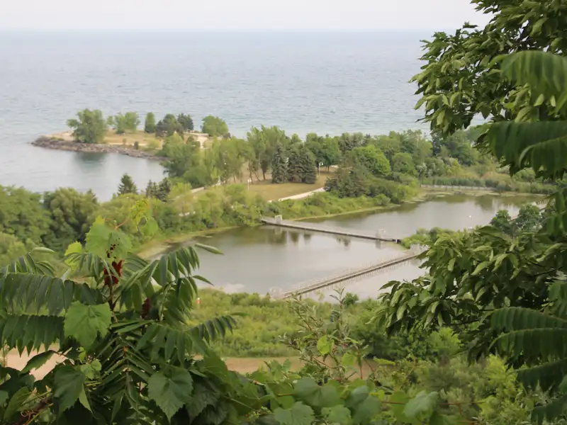Blick von einem erhöhten Aussichtspunkt auf die Küste mit einer bewachsenen Landzunge im Meer und einem See mit Stegen im Vordergrund.