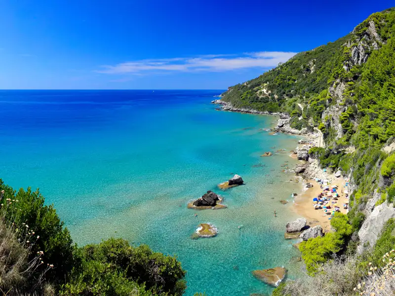 Türkisblaues Meer und Sandstrand an einer bewaldeten Küste.