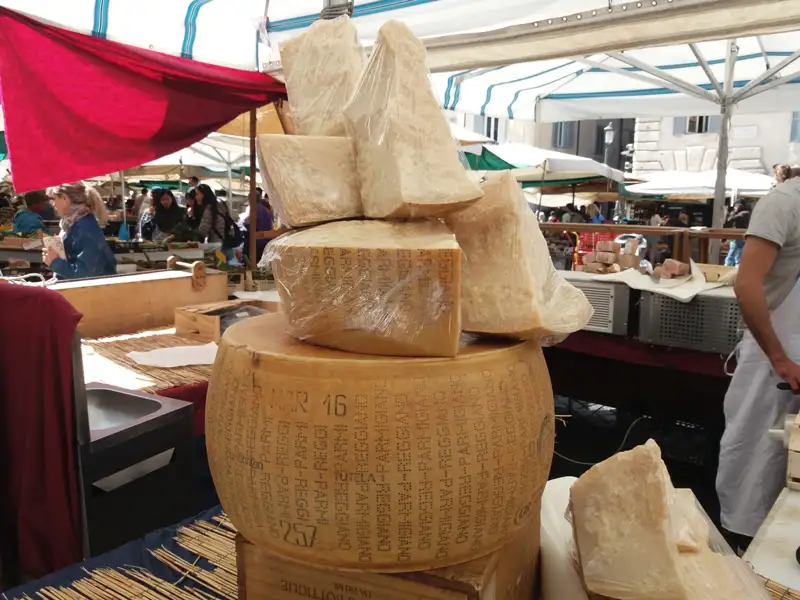 Parmesan-Käse auf einem Marktstand, möglicherweise in Parma oder Reggio Emilia, Italien. Der Käse ist in großen und kleinen Stücken zu sehen und trägt die Kennzeichnung "Parmigiano-Reggiano".
