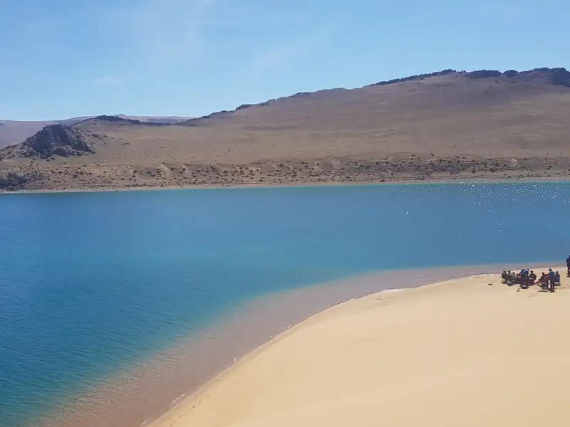 Blauer See mit Sandstrand und Personengruppe am Ufer, umgeben von kargen Hügeln.