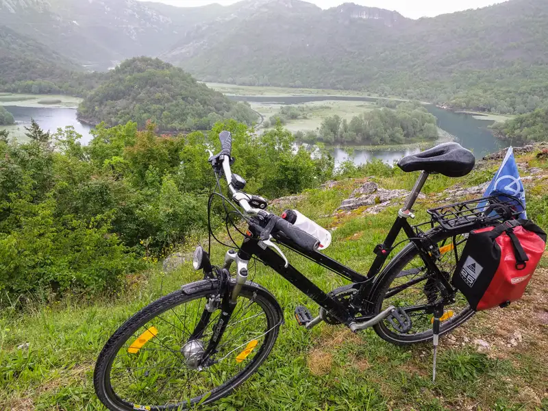 Fahrrad mit Gepäcktaschen auf einer Anhöhe, im Hintergrund ein Fluss und bewaldete Hügel.