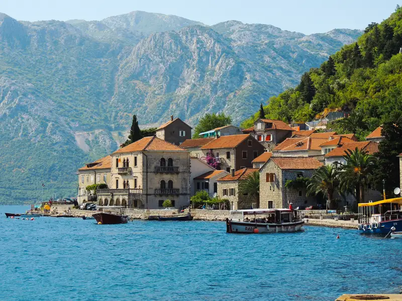 Eine malerische Küstenstadt in Montenegro, umgeben von majestätischen Bergen. Traditionelle Häuser mit roten Ziegeldächern säumen die Küste, während Boote im Hafen schaukeln.