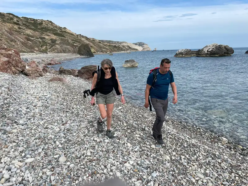 Zwei Personen wandern an einem Kiesstrand entlang, das Meer und die Küste im Hintergrund.