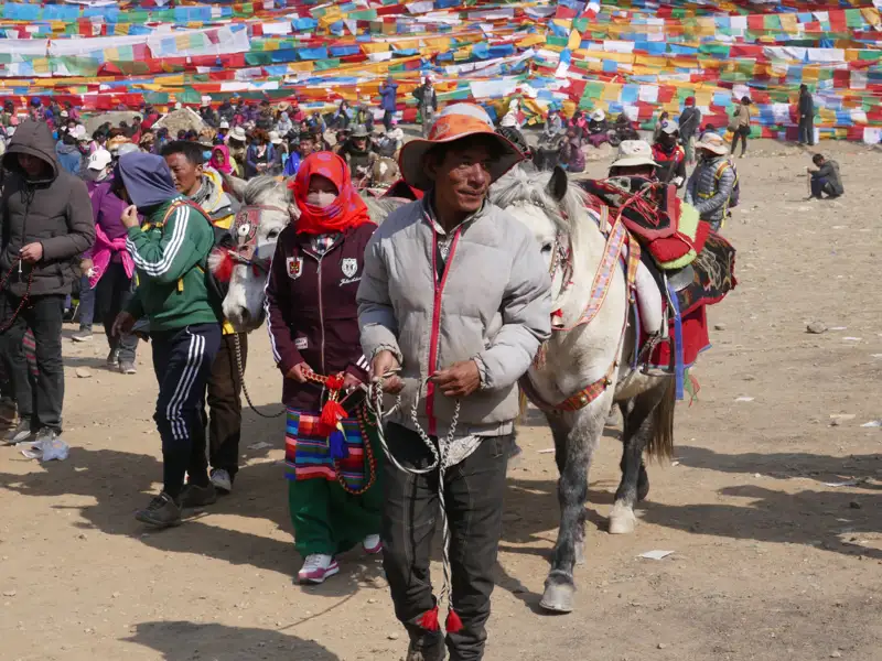 Ein Mann mit traditionellem Hut hält ein weißes Pferd mit bunten Verzierungen vor einer Wand aus Gebetsfahnen. Im Hintergrund sind weitere Menschen und Pferde zu sehen.