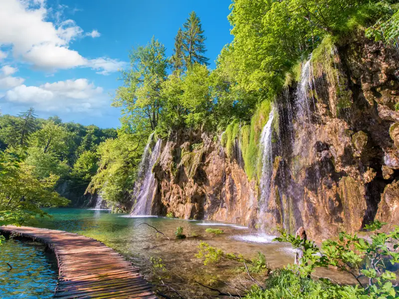 Spektakuläre Wasserfälle, die über eine Felswand in einen türkisfarbenen See im Nationalpark kaskadieren. Ein Holzsteg schlängelt sich durch das flache Wasser und bietet Zugang zu dieser atemberaubenden Naturkulisse.