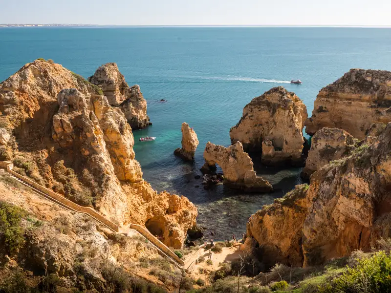 Spektakuläre Felsformationen an der Küste, ein Boot fährt durch das türkisblaue Wasser, eine Treppe ermöglicht den Zugang zum Strand.