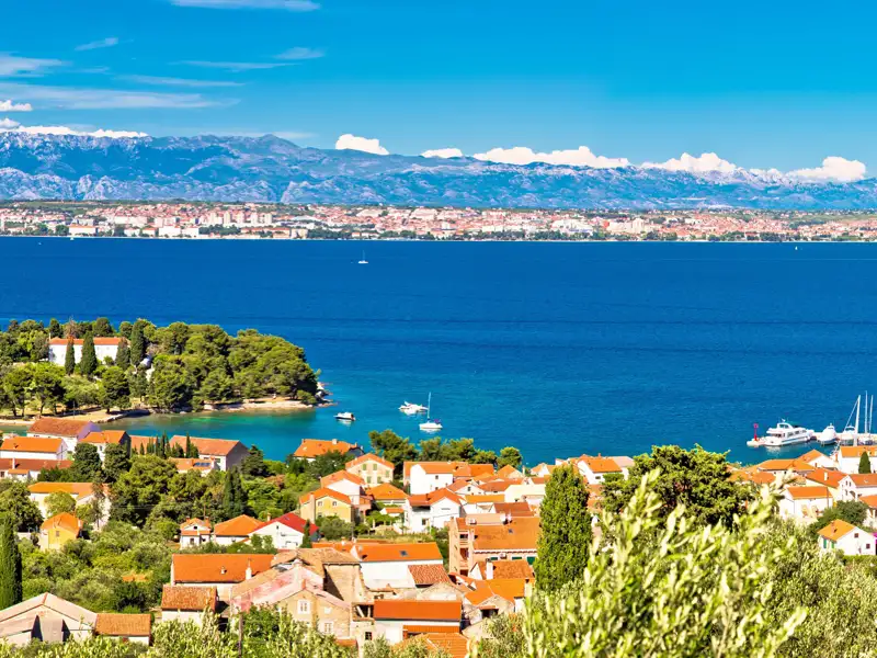 Idyllischer Blick auf eine Küstenstadt mit roten Dächern und türkisfarbenem Wasser. Im Hintergrund erstreckt sich eine Bergkette.