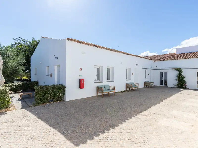 Weiße Unterkunft mit gepflastertem Vorplatz und Sitzgelegenheiten.