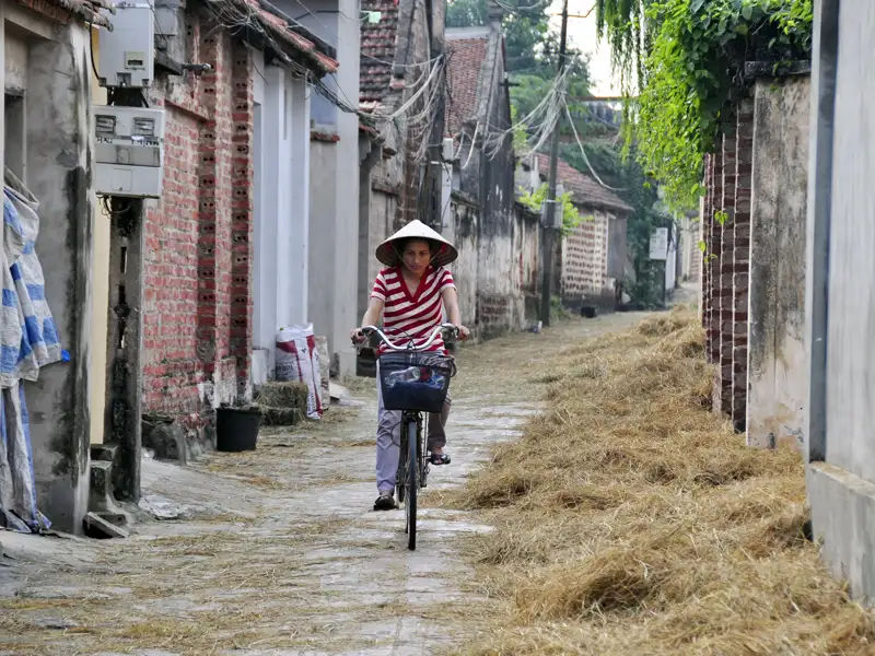 Person mit traditionellem Hut fährt Fahrrad durch Dorfgasse.
