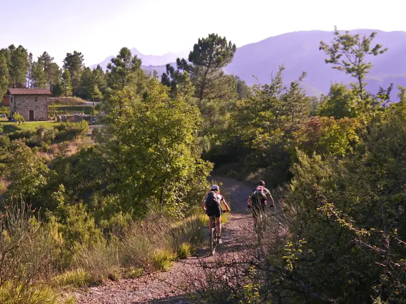Zwei Mountainbiker auf einem Bergpfad, umgeben von Vegetation. Ein Gebäude und Berge sind im Hintergrund sichtbar.
