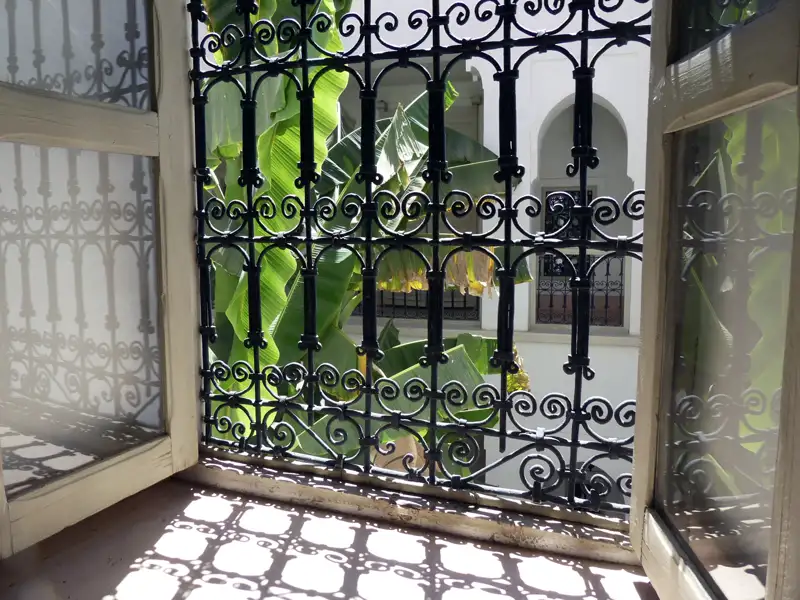 Blick durch ein traditionelles marokkanisches Fenstergitter auf einen Innenhof mit Bananenpflanzen.